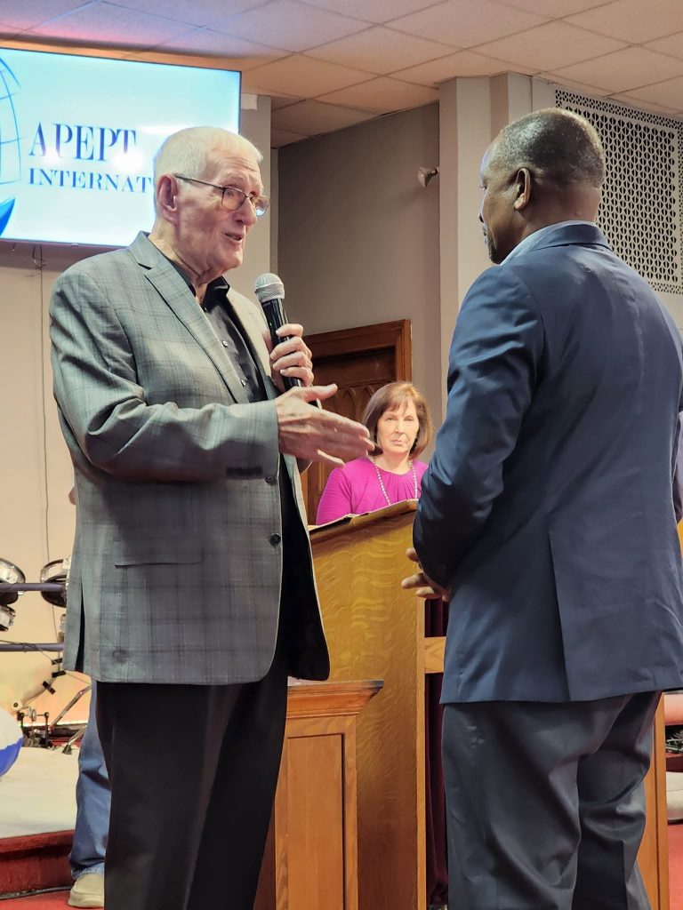 A speaker holding a microphone addresses another man beside a church lectern