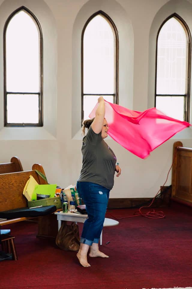 A woman holding a bright pink worship flag in a church sanctuary with arched windows
